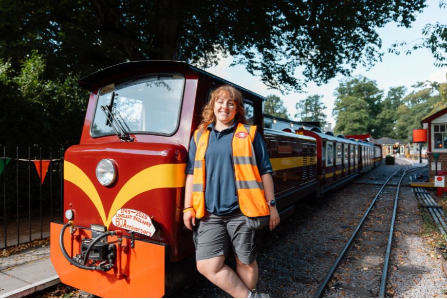 A woman in an orange high vis jacket standing next to a little red train